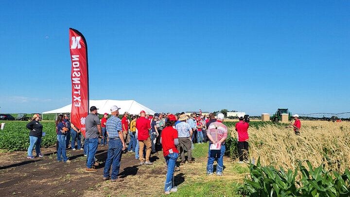 Group of people gathered in a farm field under a clear blue sky, near a red Extension banner and a white tent.