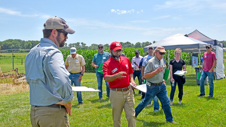 Group of people discussing in a sunny farm setting with crops and tents visible.