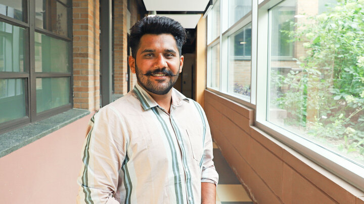 Akashdeep Kamboj poses for a photo in a hallway lined with windows on both sides. He his wearing a button-up shirt with peach and green vertical stripes.