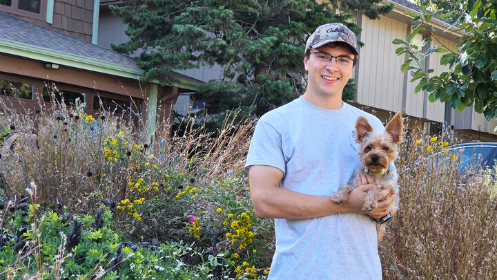 Man holding small dog in front of a house with garden and greenery.