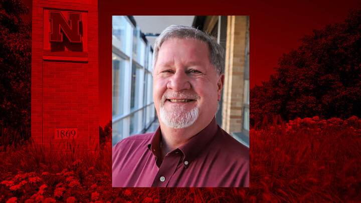 Man in a red button up shirt in front of a red background with a University of Nebraska emblem.