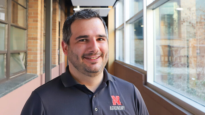 Man wearing a black polo shirt with red "N Agronomy" text, standing in a corridor with large windows.