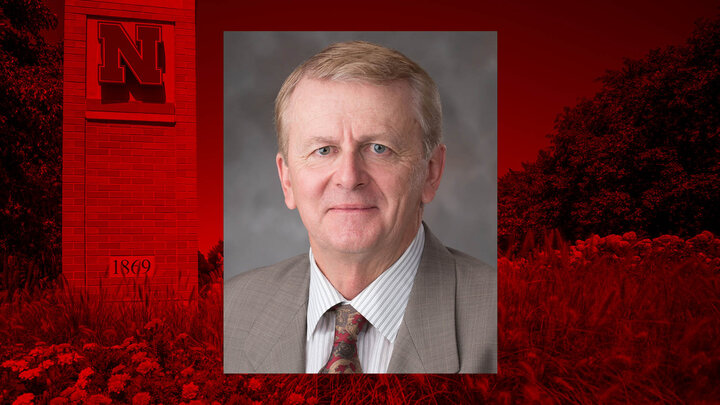 Man in a suit and tie against a red background with a University of Nebraska emblem.