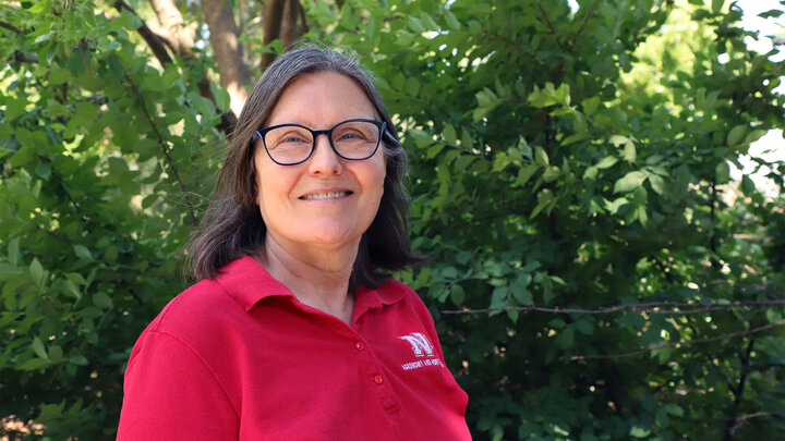 Woman in red polo shirt standing in front of lush green foliage outdoors.