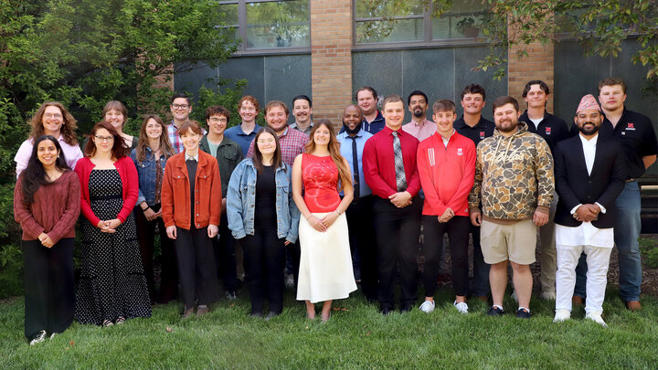 Group of people posing outdoors in formal and casual attire, with greenery and a building behind.