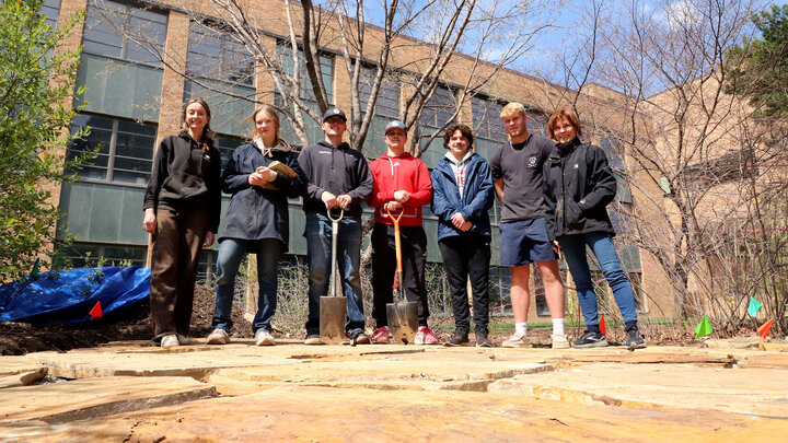 Group of people outdoors with shovels, standing in front of a brick building and bare trees.