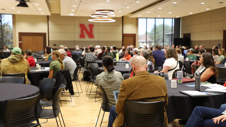 Conference attendees sit around circular tables in a large ballroom in UNL's East Campus Union. A prominent red N stands out on the otherwise blank back wall.