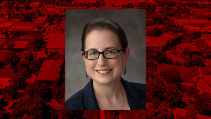A woman with her hair pulled back in a blazer, with a red-filtered aerial view of UNL's east campus in the background.