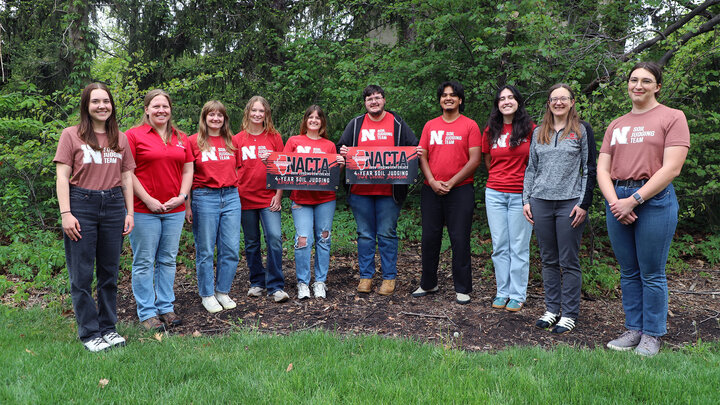 Group of people standing outdoors, some wearing red shirts with logos, holding signs, surrounded by greenery.