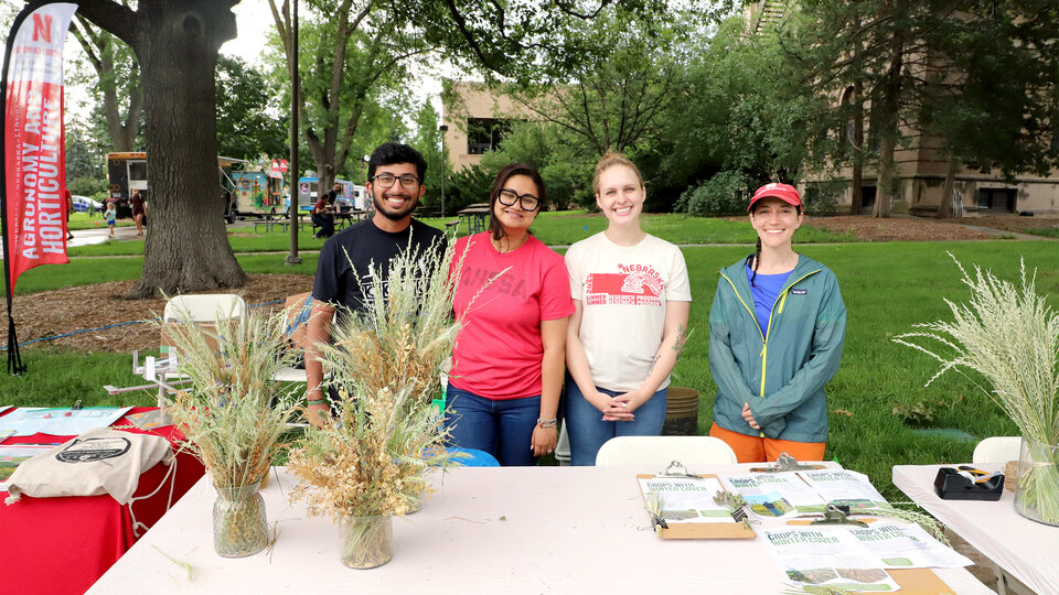 Four people smiling behind a table with plants and brochures, outdoors in a park setting.