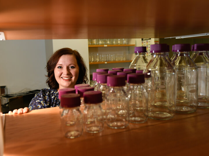 Allison Barnes smiles at the camera, posing behind a shelf with different sized vials and bottles on it