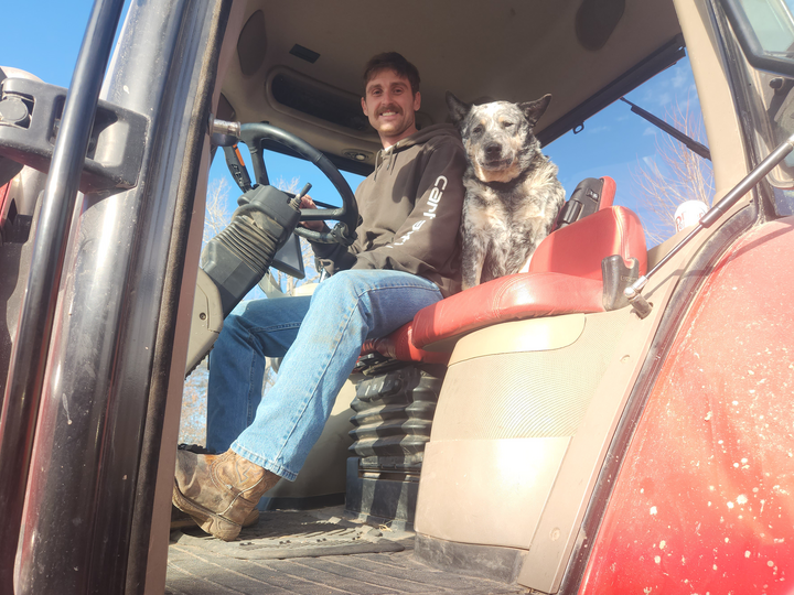 Justin Zoucha smiles at the camera from inside a red tractor, a cattle dog sitting to his left