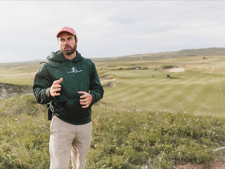 A candid photo of Andrew Getty standing in a golf course at The Prairie Club, a large expanse of manicured golf course turf behind him