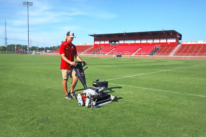 Groundskeeper mowing a green sports field near red stadium seats under a clear blue sky.