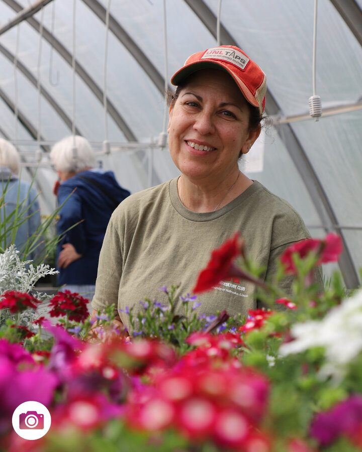 Woman in ball cap stands in greenhouse surrounded by flowers.