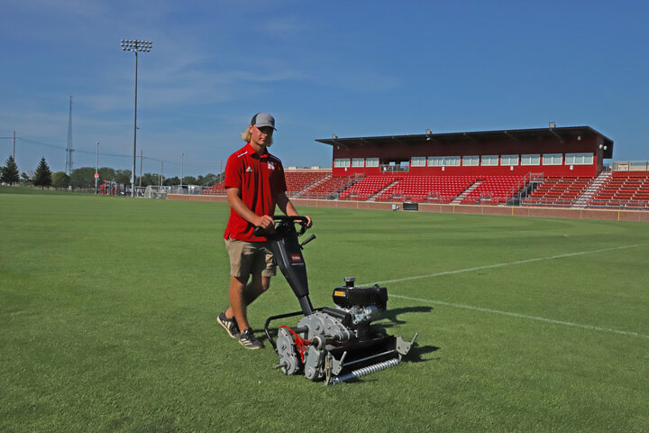 Groundskeeper mowing a green sports field near red stadium seats under a clear blue sky.